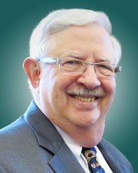 Smiling elderly man in a suit, wearing glasses, with a light background. The image conveys professionalism and friendliness, relevant for a biography or profile.