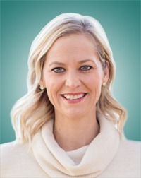 Smiling woman with long, wavy blonde hair in a cream sweater, set against a soft green background, conveying a warm and approachable demeanor.